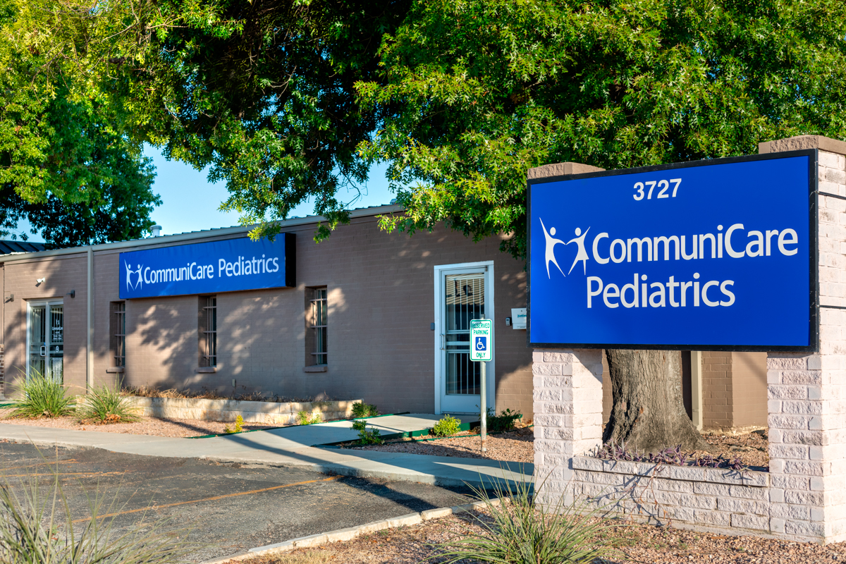Exterior view of the CommuniCare Roosevelt Campus pediatric clinic in San Antonio, Texas, showing the building’s pale brown brick facade, prominent blue signage with white text that reads "CommuniCare Pediatrics," and surrounding trees and shrubbery.