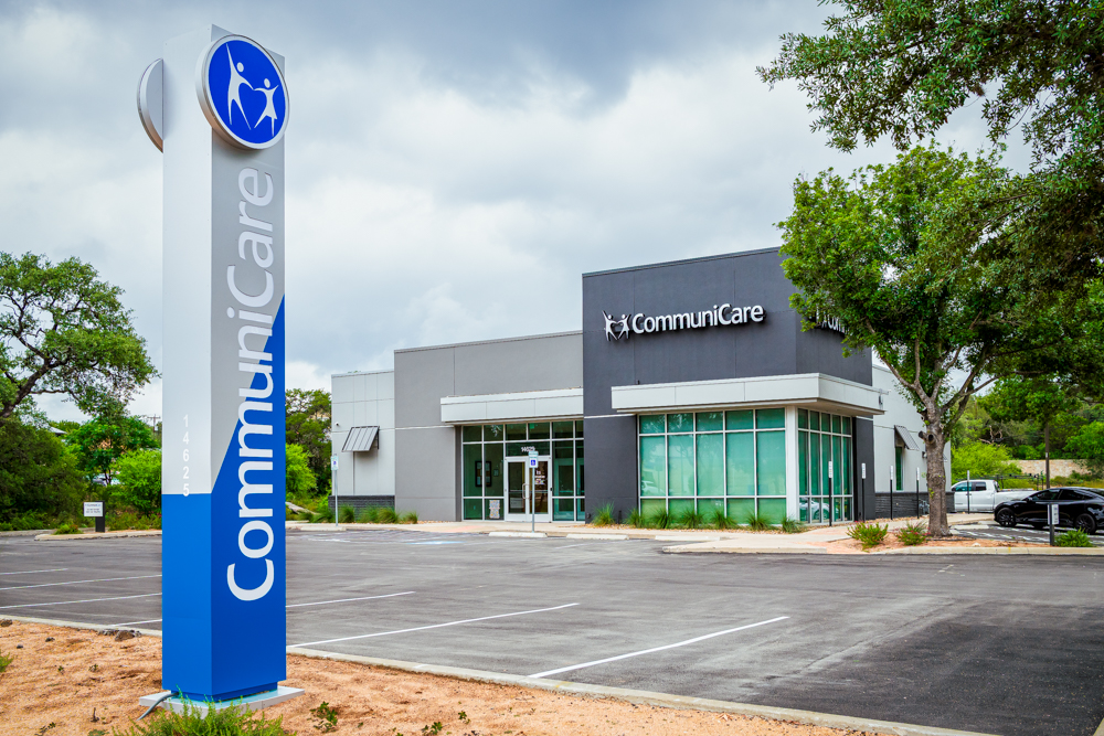 A modern CommuniCare building with a gray and white exterior, large front windows, and the CommuniCare logo on the facade. A tall blue and white sign in the foreground displays the name 'CommuniCare' vertically. The parking lot is empty, and the scene is framed by green trees under a cloudy sky.