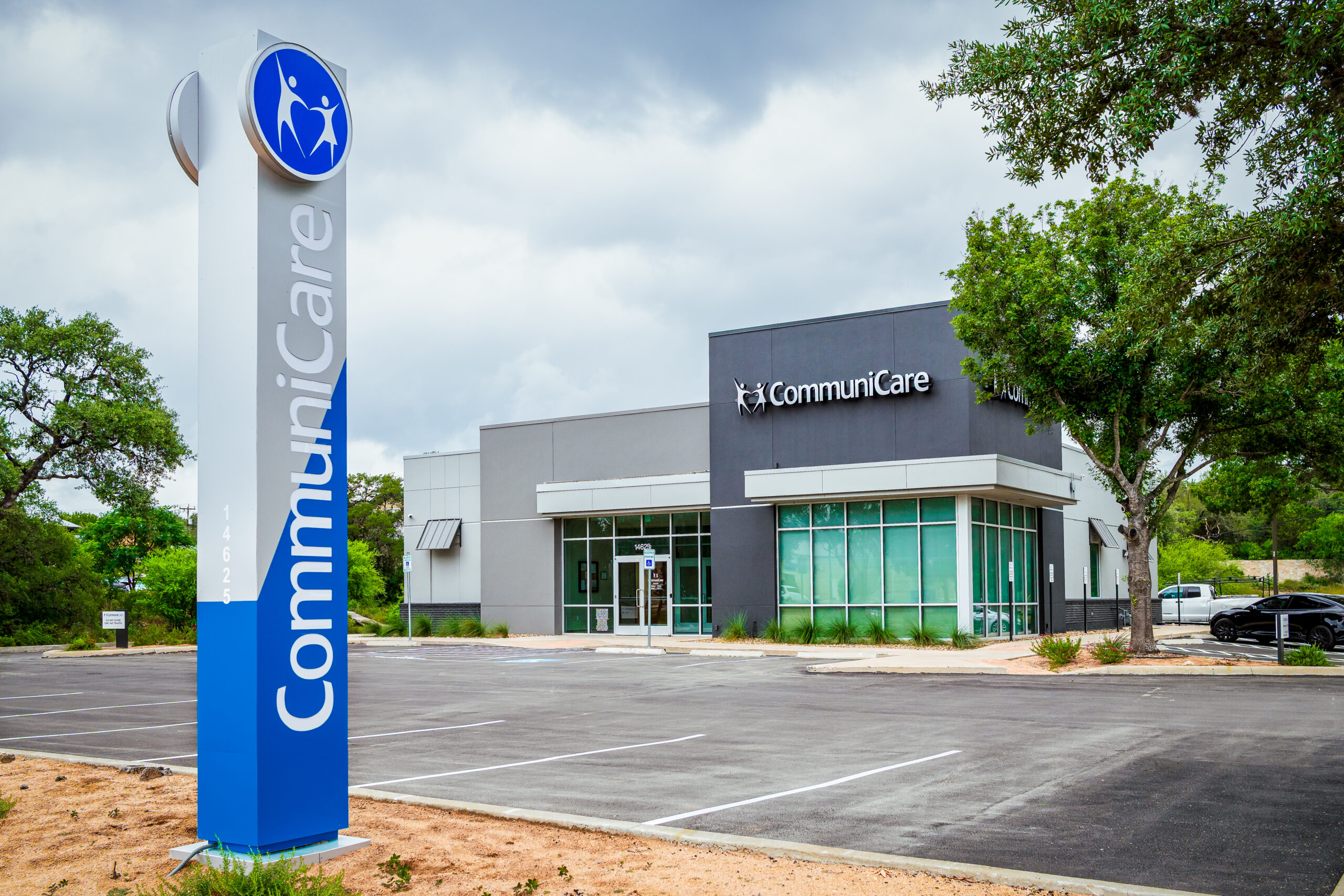 A modern CommuniCare building with a gray and white exterior, large front windows, and the CommuniCare logo on the facade. A tall blue and white sign in the foreground displays the name 'CommuniCare' vertically. The parking lot is empty, and the scene is framed by green trees under a cloudy sky.
